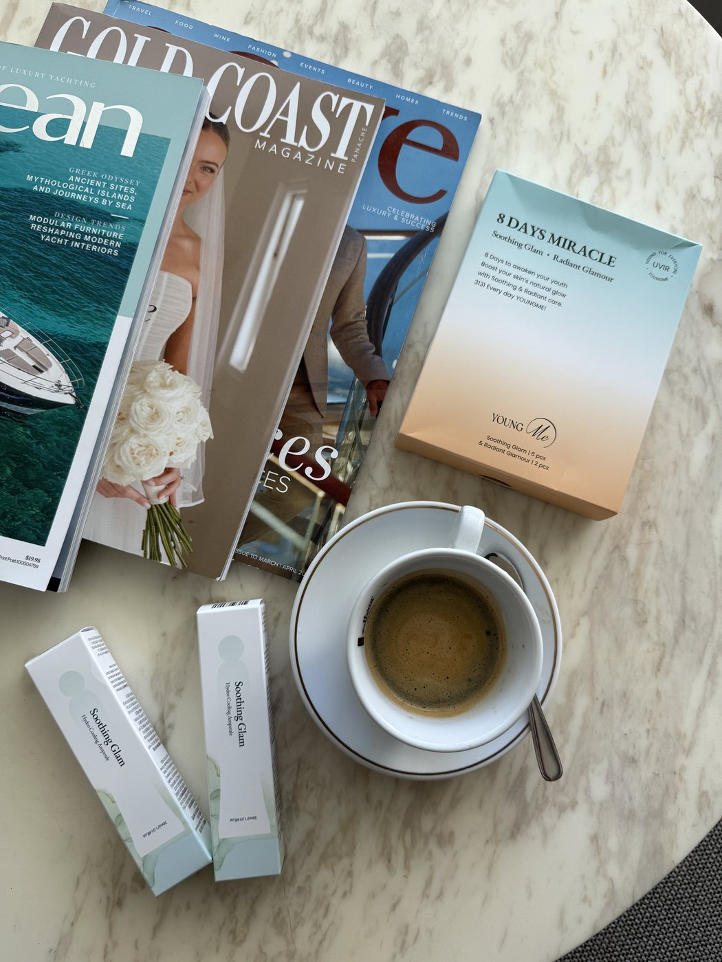 Flat lay of magazines, skincare boxes and a coffee on a marble table