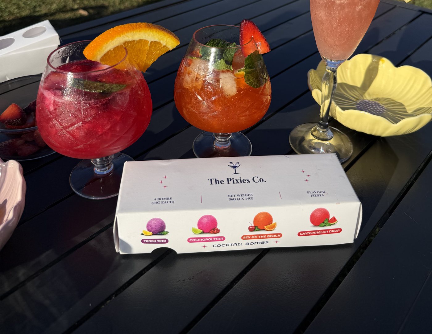 Three fruity cocktails on a garden table beside a box of cocktail kits, Easter eggs on the lawn behind